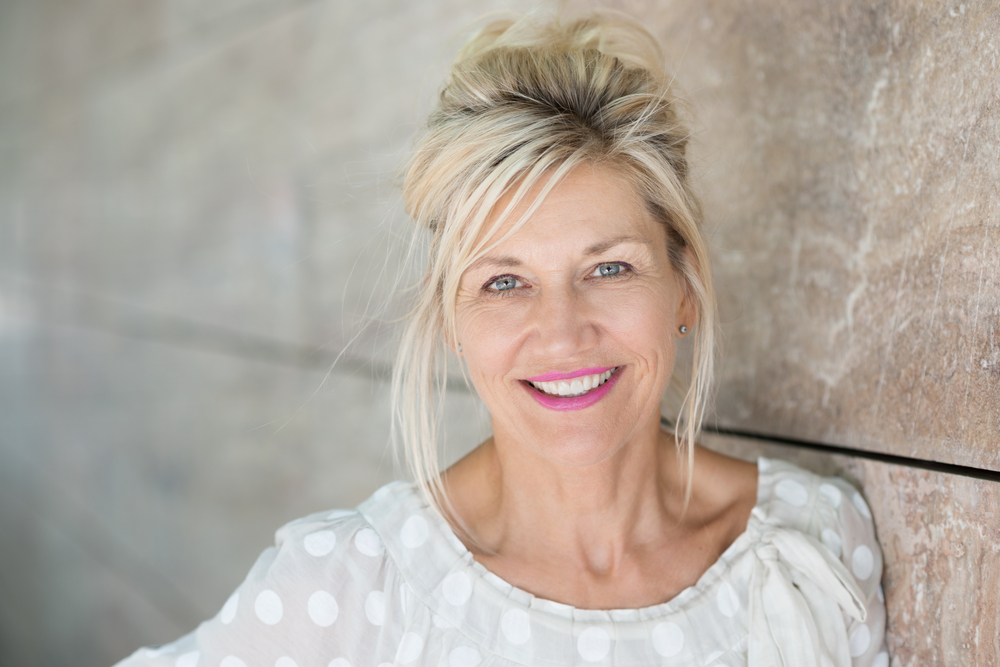 A smiling blonde woman with bright pink lipstick leans against a textured wall at Mountainview Periodontics